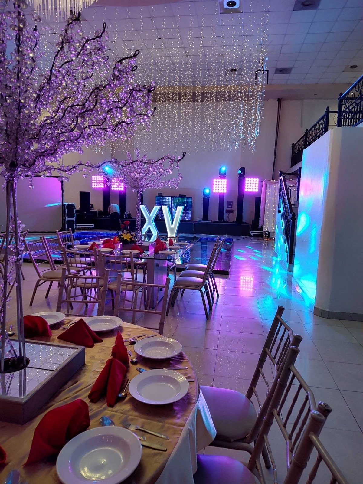 Mesa montada con vajilla, servilletas rojas y árboles iluminados frente a la pista de XV años en VillaMont